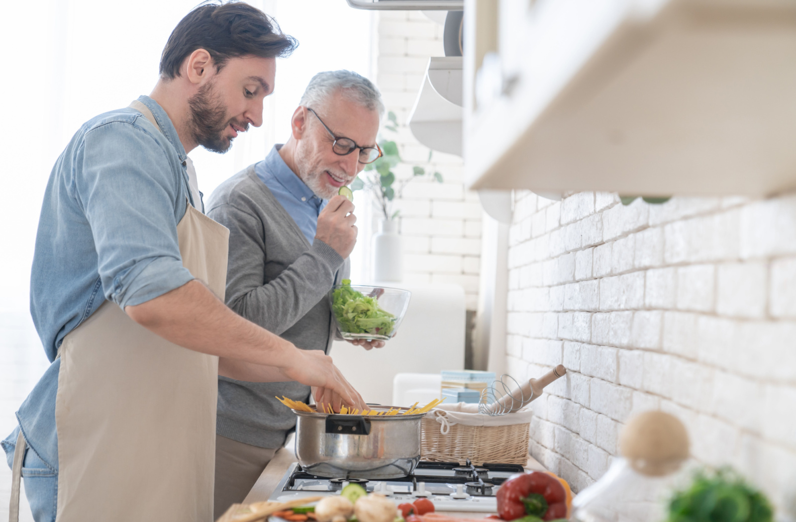 A young man helping his senior father cook past in their brightly lit kitchen.