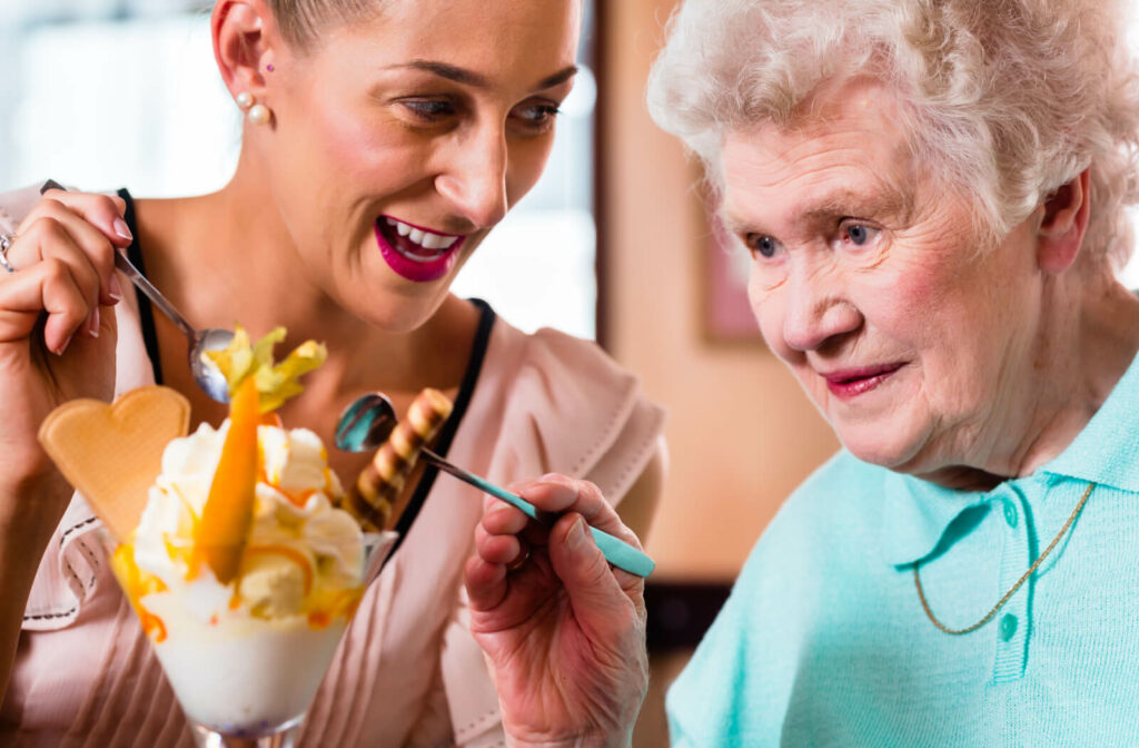 A grown daughter is enjoying an ice cream sundae with her elderly mother, who has dementia, creating joyful moments together.