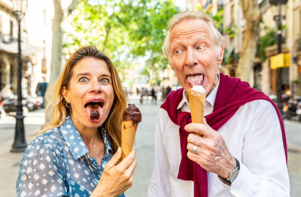 A grown daughter shares a playful moment with her elderly father, who has dementia, as they enjoy a cone of ice cream together on the street.