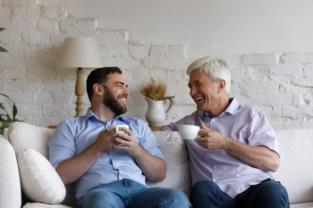 A senior father and his son happily chatting on the couch & drinking coffee