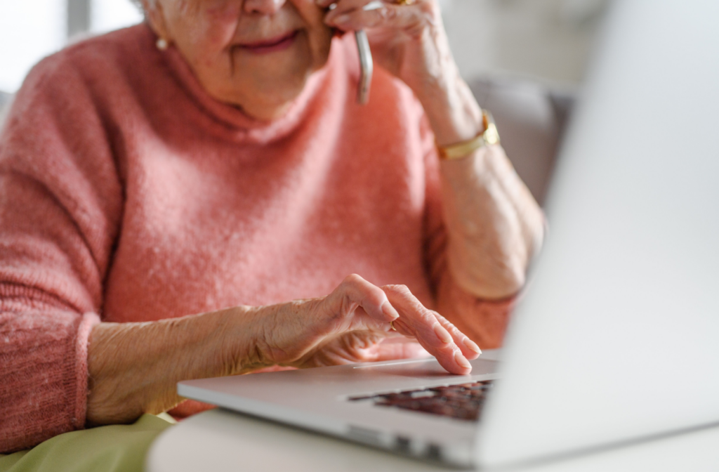 A senior sits at their laptop with a phone to their ear and fingers scrolling the trackpad.