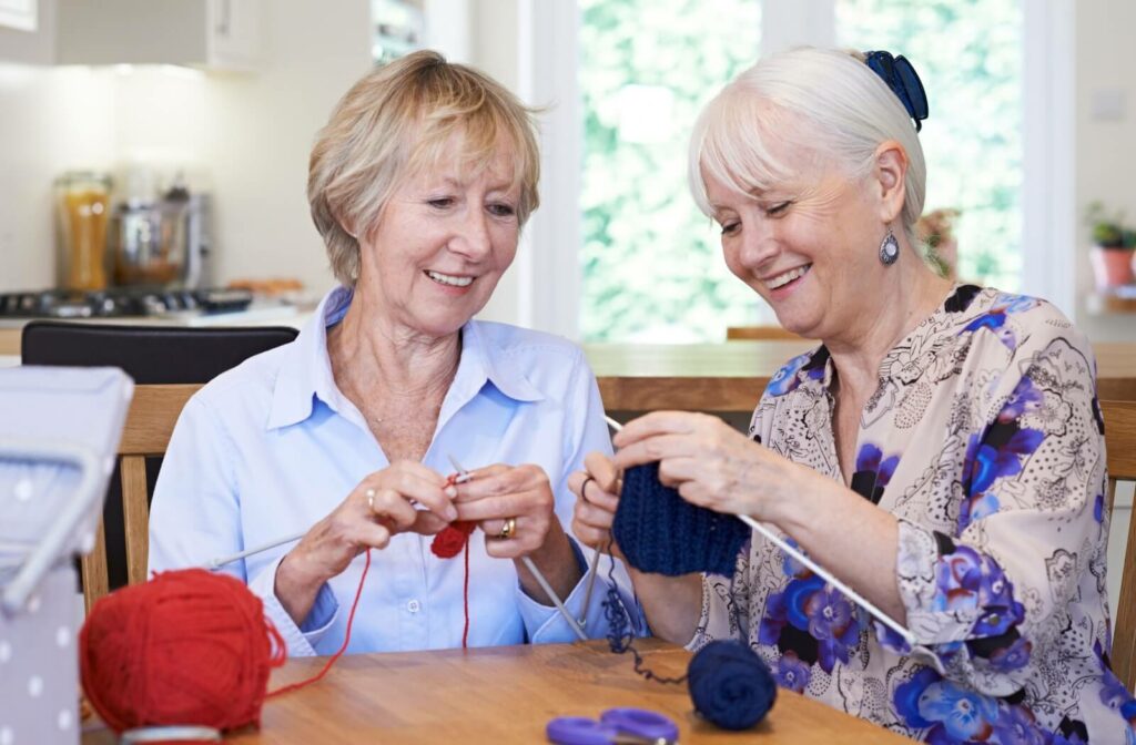 Two seniors sit at a dining room table, helping each other begin knitting projects using red and blue yarn