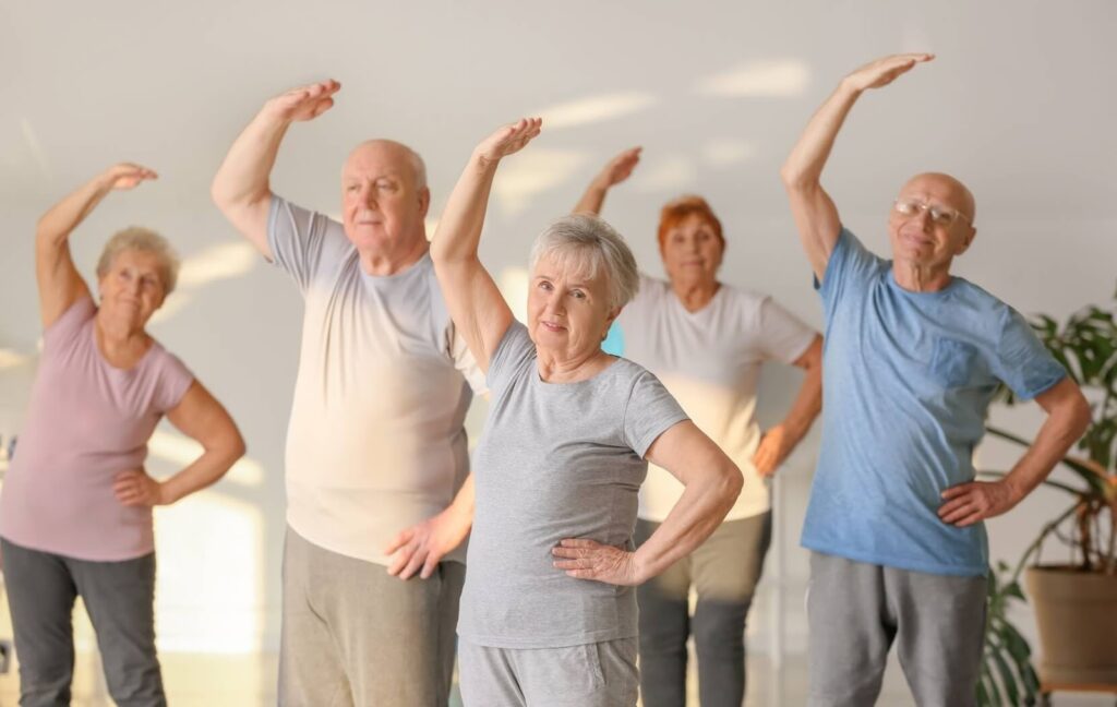 A group of seniors wearing workout clothes raise their arms over their heads to stretch before a group exercise class.