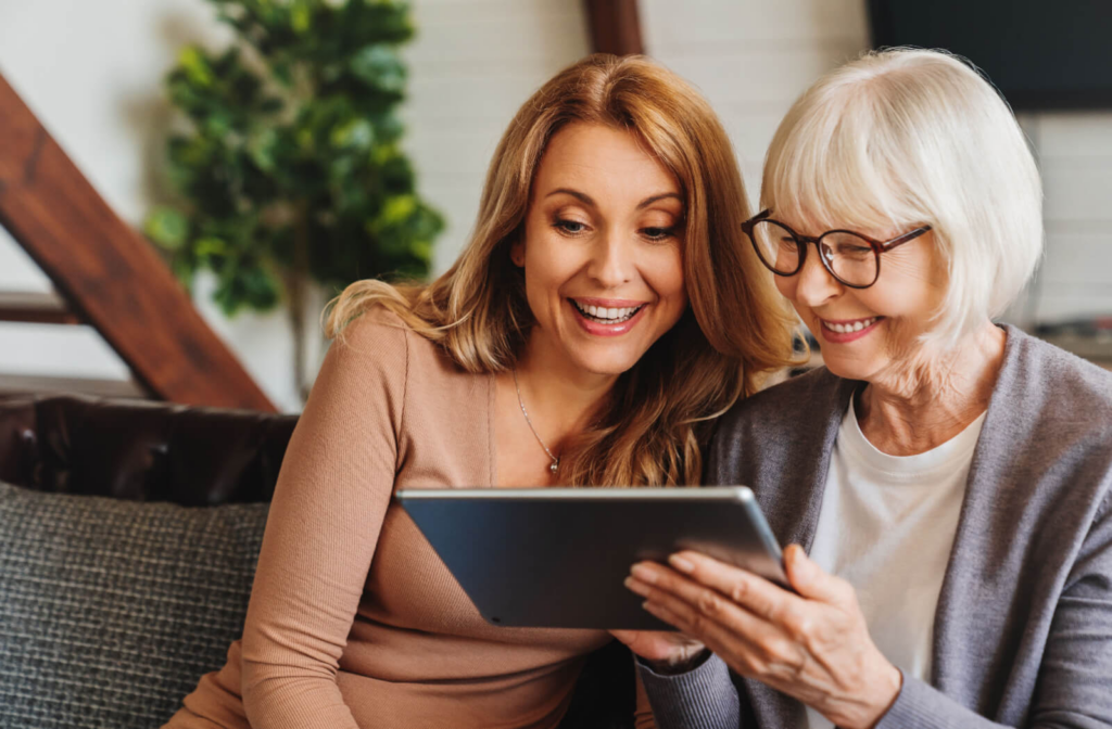 An adult child smiles while their older parent holds up a tablet to show them pictures of a potential assisted living community