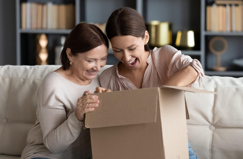 An adult child and their older parent reach into a box while sitting on the couch and laughing in a new home in assisted living