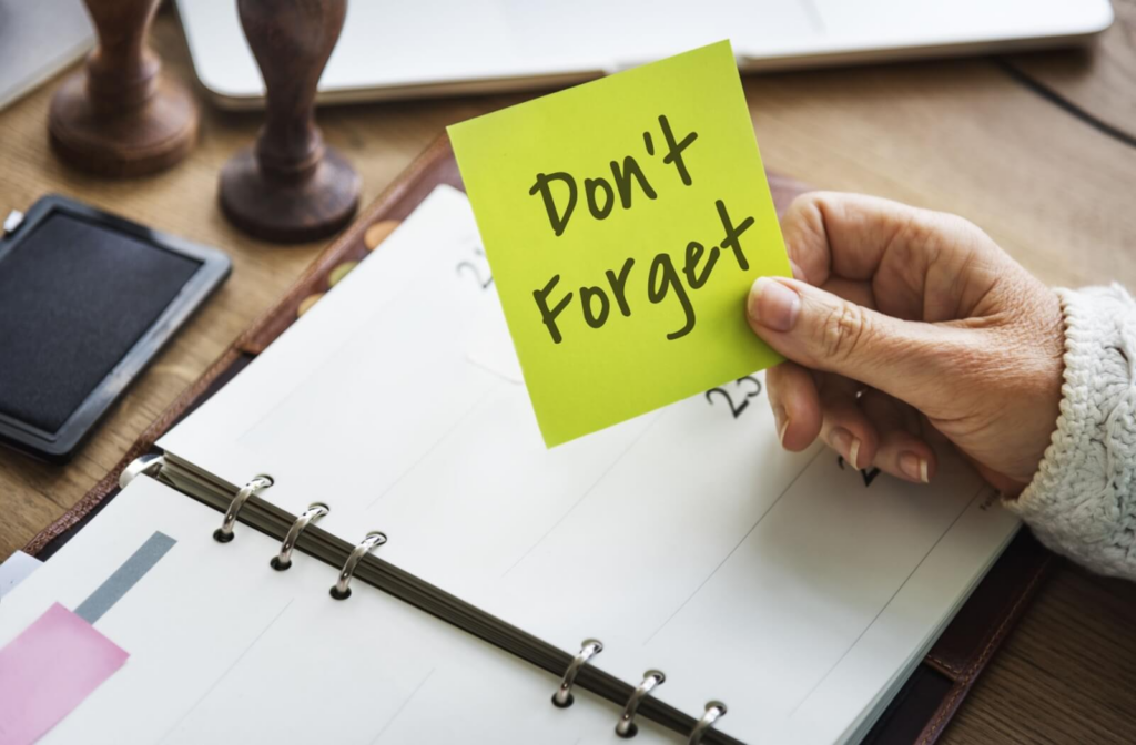 An overhead shot of a cluttered desk and an older adult holding a yellow note saying "Don't forget"