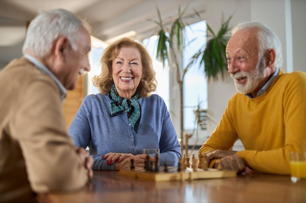 two seniors play chess while a third looks on smiling