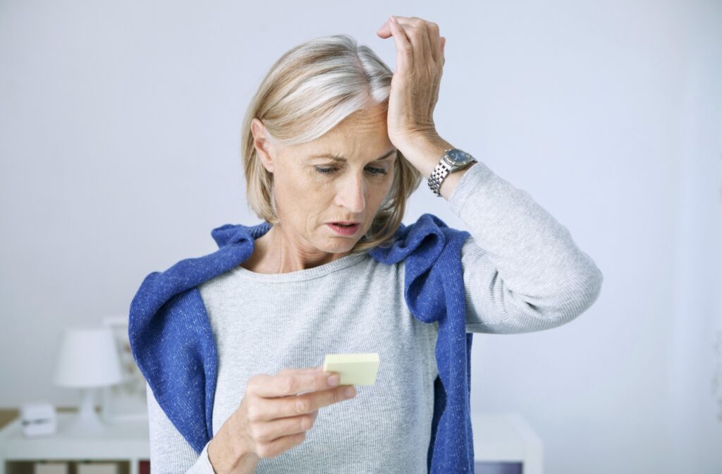 An older adult rubs their forehead in confusion while reading a small post-it note they've left themselves